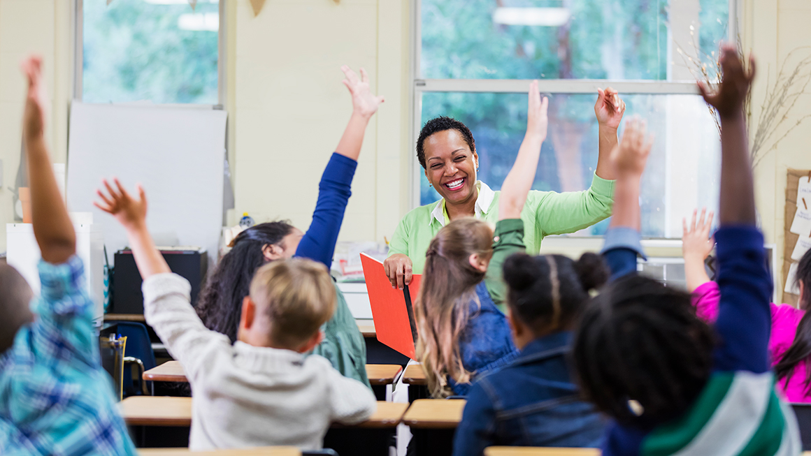 An African-American teacher sitting in front of her class of elementary school students with most of the children are raising their hands. 