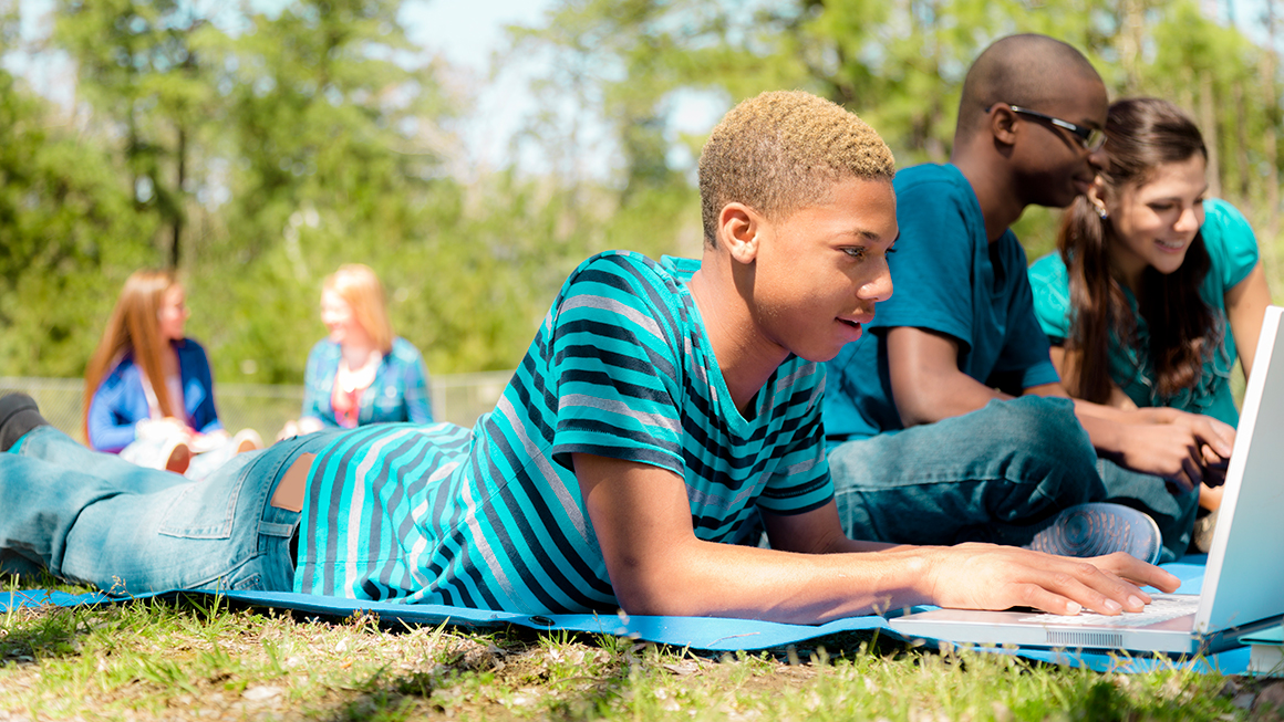 Multi-ethnic group of high school friends hang out on campus or park. Studying with laptop, books.