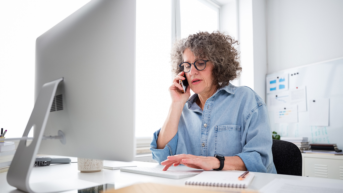 Middle-aged woman with curly gray hair wearing glasses and a denim shirt, talking on the phone while working at a desktop computer in a bright, modern office.